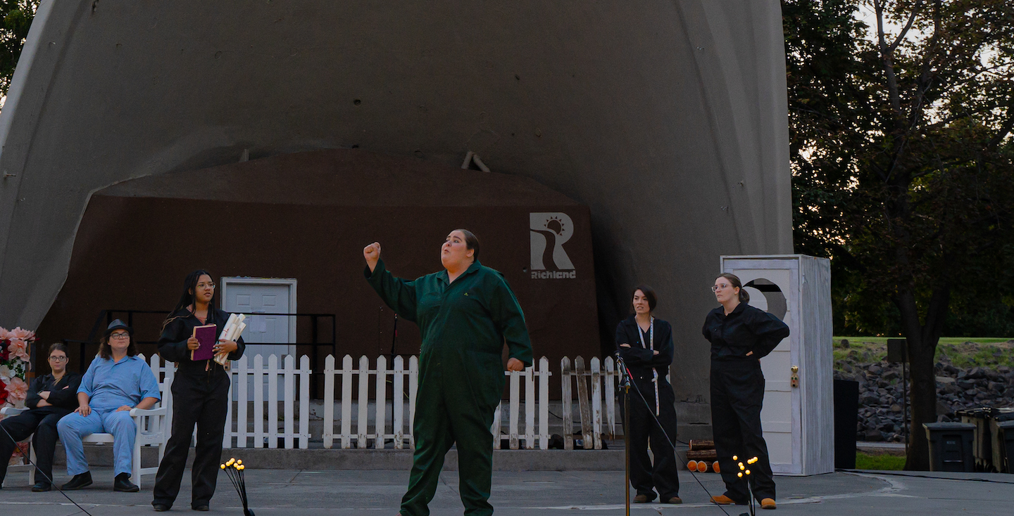 A group of people in a an outdoor theater, mid-performance. A large arch-like concrete structures rest behind the actors. In front of that structure is a white wooden fence and a white door off to the right. Two actors can be seen on the left sitting in chairs. Anothor holding papers and a book standing to the left of the actor in the center of the image. The actor in the center has a green jumpsuit on and their right hand raised gesturing towards something. Two more actors are standing to the right of the center actor in the background. All actors are looking at the one in the center.