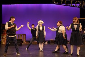 A group of actors spread out on a theater stage. All of the performers are very animated, and several are playfully gesturing towards the performer in the center of the stage.