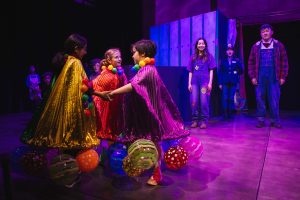 A group of youth dressed in costumes holding hands in a circle on a colorfully lit theater stage.