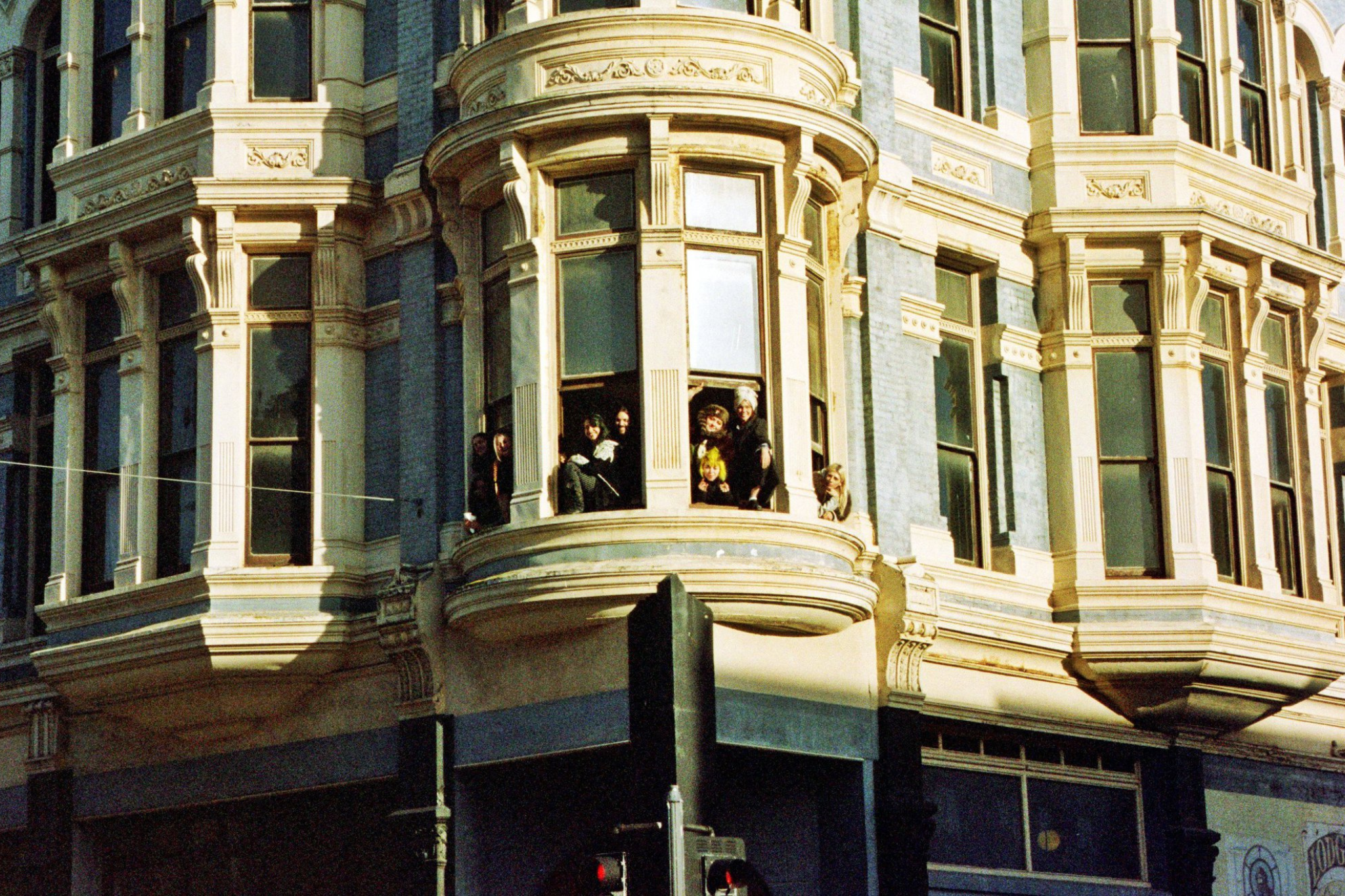 An ornate opal building lit by evening sunlight shows several people peering out of the winders to see what may be going on in the streets below them.