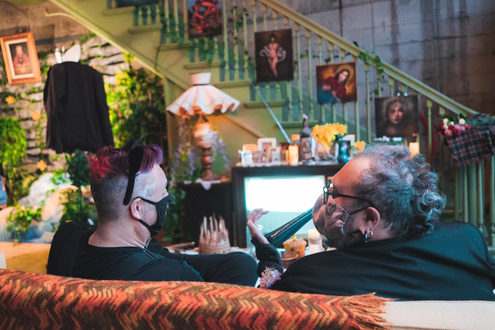 Image shows two people wearing masks around the nose and mouth. We are watching them have a conversation on a couch and colorful room all from behind..