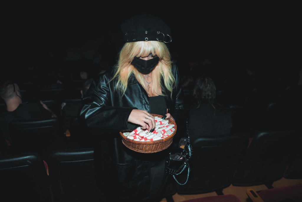 One person with shoulder-length blonde hair wearing all black with a black hat and mask. They are reaching into a bowl of scrolls.