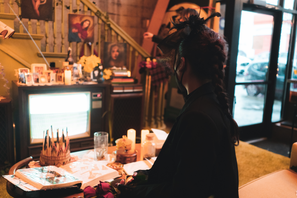 A person sits with their back towards the camera wearing all black. they are positioned in front of a table with various books, candles, and cups, and TV playing static feedback.