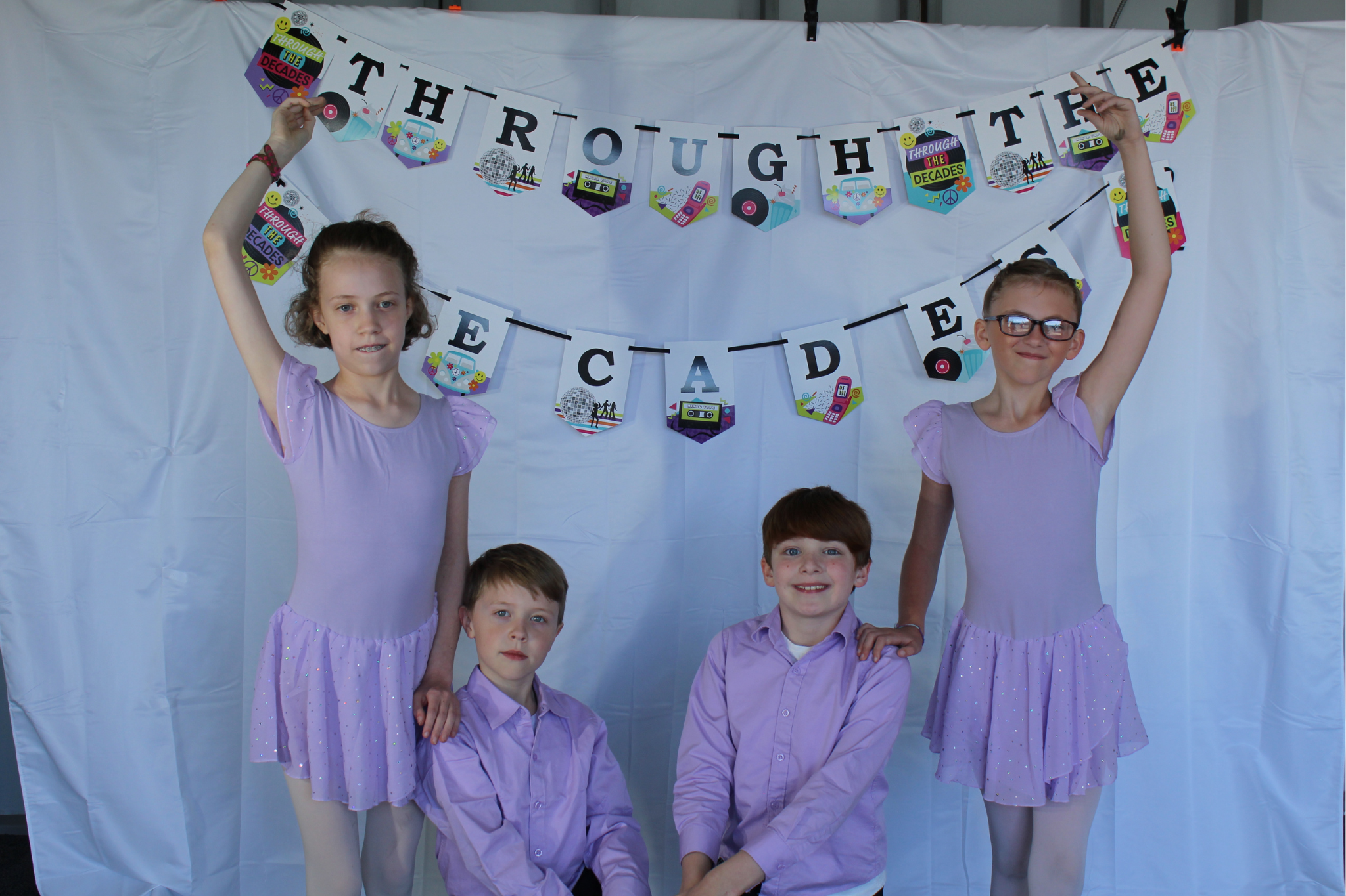 Young dancers posing in their purple ballet costumes against a white backdrop that says, "Through the Decades".