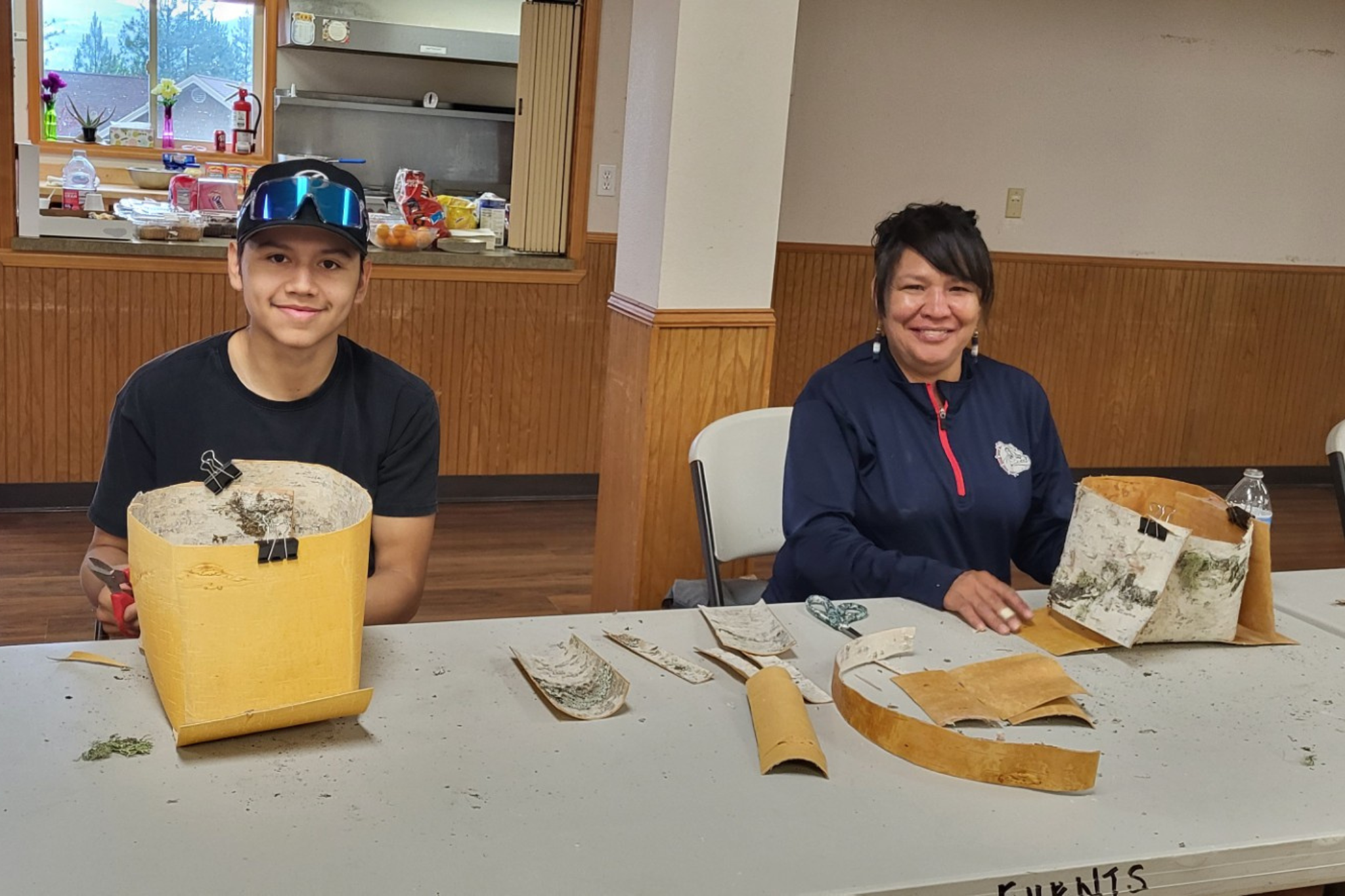 Two people smiling at a camera in the middle of crafting a woven basket.