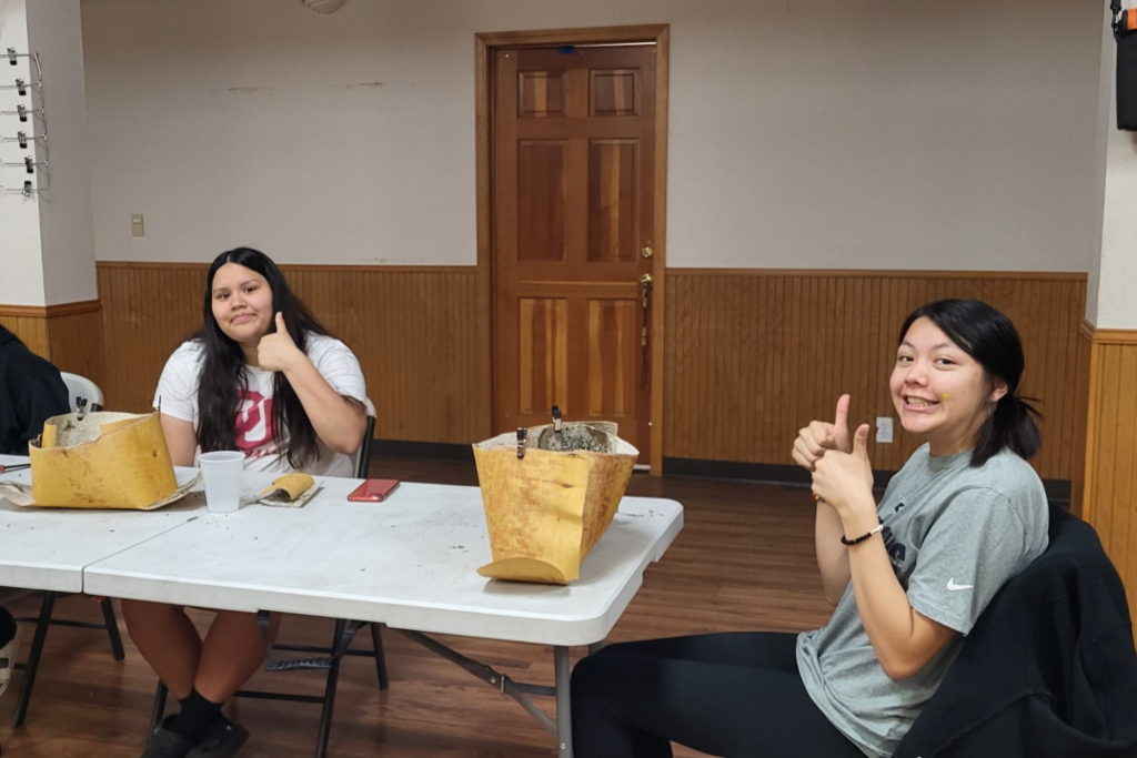 Two young people sitting at a table smiling with their thumbs up, while working on a project.