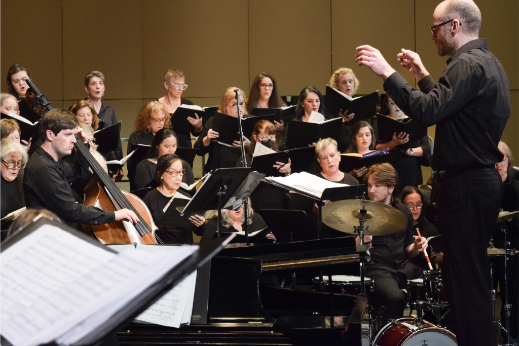 A group of people dressed in black singing. A conductor standing before the choir.